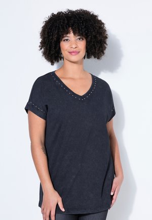 Kiharatukkainen nainen, joka pukeutunut tummanväriseen V-pääntieiseen lyhythihaiseen paitaan, jossa on pieniä niittejä pääntien reunoilla, seisoo valkoista taustaa vasten.