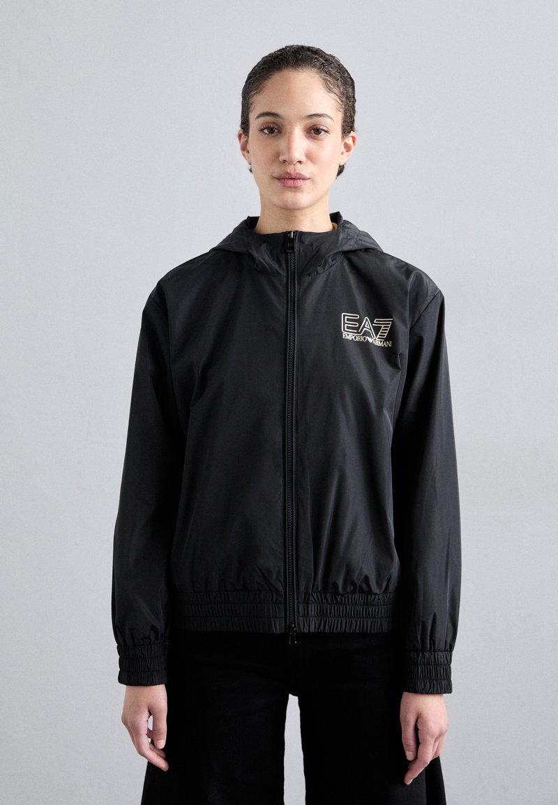 Casaco leve preto com capuz, com punhos e cintura elásticos, apresentando um logotipo EA7 bordado em dourado no peito. Textura suave.