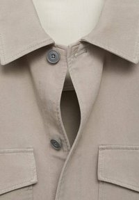 Beige collared shirt with two front pockets, gray buttons, and a plain fabric texture. The neckline features a shallow opening.