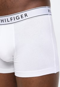 Gros plan sur une personne portant un boxer blanc Tommy Hilfiger avec une ceinture de marque sur une peau foncée.