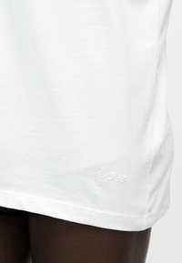 Detailný záber bielej látky s vyšitým nápisom „BOSS“ pri okraji, nosenej na tmavej pokožke.