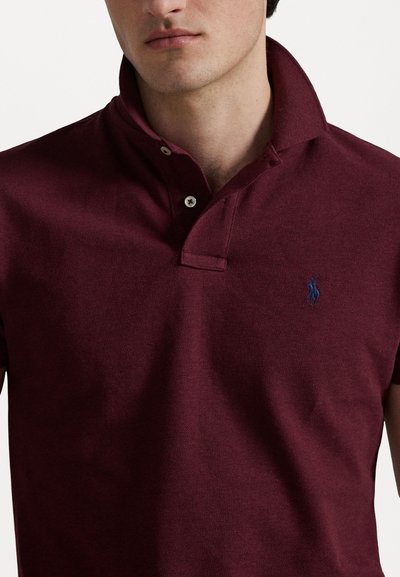 Polo bordeaux en tissu texturé, doté d'un col boutonné et d'un petit logo bleu sur la poitrine gauche.