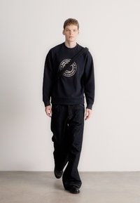 Tumši zils sporta krekls ar apaļu logotipu centrā, kopā ar vaļīgām tumšām džinsu biksēm un melnām kurpēm. Vienkāršs, ikdienišķs apģērbs.