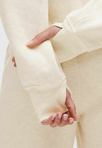Sweatshirt u boji kreme s rebrastim manžetama i mekom teksturom, na slici se vidi ruka koja drži rukav. Tkanina izgleda lagano i ležerno.