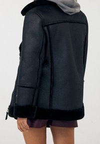 Giacca in shearling nera con collo rotondo, superficie testurizzata, maniche lunghe e un soffice bordo nero all'orlo. Design semplice e moderno.