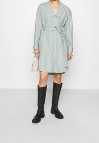 Světle zelené šaty na knoflíky s velkým límcem, páskem v oblasti pasu a dlouhými rukávy, doplněné černými koženými botami po kolena.