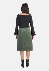 Falda midi de pana verde con corte recto, que presenta dos bolsillos traseros y una etiqueta en la cinturilla. Combinada con una camiseta negra de mangas largas.