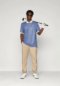 Μπλε και λευκή ριγέ πόλο μπλούζα με γιακά, μπεζ παντελόνι και λευκά αθλητικά παπούτσια. Το άτομο κρατάει ένα γκολφ με ένα γάντι σε ένα χέρι.