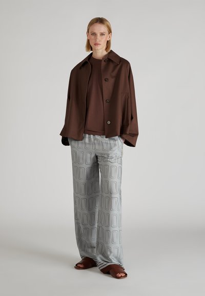 Brązowa, oversize'owa kurtka z guzikami, zestawiona z brązową koszulą i jasnoszarymi, wzorzystymi spodniami typu wide-leg. Brązowe klapki dopełniają stylizację.