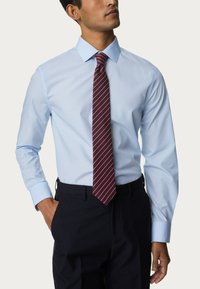 Camicia a maniche lunghe blu chiaro in tessuto liscio, abbinata a una cravatta a righe bordeaux. L'outfit è completato da pantaloni scuri.