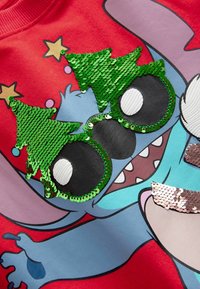 Felpa rossa con applicazioni di paillettes a forma di alberi di Natale verdi e grandi occhiali in stile cartone animato su un'illustrazione di un personaggio blu.