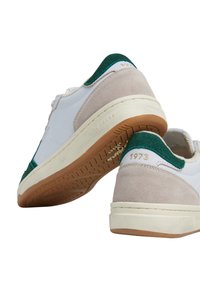 Bela teniska z zelenimi in bež akcenti; gladka usnjena in semenska tekstura, z gumijastim podplatom in ožigosanim detajlom '1973'.