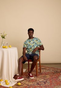 Heren overhemd met korte mouwen in een blauw-groen bloemenpatroon, gecombineerd met marineblauwe shorts en rode sneakers. Eenvoudige houten stoel en tafel met citroenen.