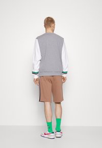 Man som bär en grå ärmlös sweatshirt, en vit långärmad tröja, bruna shorts, gröna strumpor och vita Nike sneakers, står vänd bort.