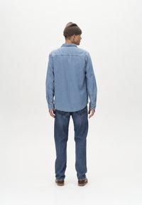 Világoskék farmering hosszú ujjakkal, gombos mandzsettával, ívelt hemvonalával, sötétkék farmernadrág felett viselve, barna kötött sapkával.