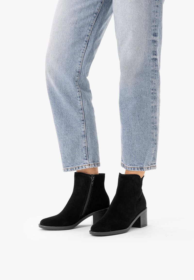 Femme portant un jean bleu clair et des bottines en daim noir à talons blocs avec fermetures éclair latérales sur fond blanc.