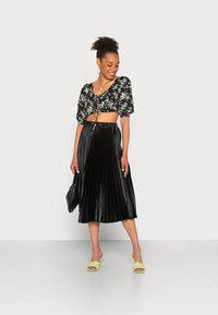 Top curto preto com padrão floral e mangas bufantes, combinado com uma saia midi preta plissada. Os acessórios incluem uma bolsa preta e sandálias de salto bloco verde claro.