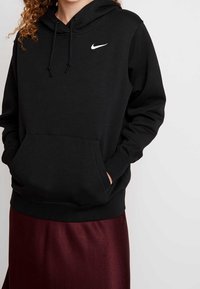 Kvinna i svart Nike-hoodie med framficka och burgundy kjol, händer delvis i fickorna, lockigt hår synligt i övre vänstra hörnet.