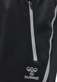 Black athletic pants with a grey zippered pocket, feature a white stripe along the side, and a logo printed near the hem. Smooth texture.