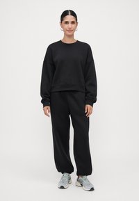Žena s vlasy svázanými do těsného drdolu, oblečená v černé oversized mikině a kalhotách s světle šedými teniskami, stojící proti čistě bílemu pozadí.