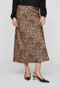 Nő leopárdmintás midi szoknyában, fekete hosszú ujjú blúzban és fekete lakk slingback sarkú cipőben, egy szürke háttér előtt áll.