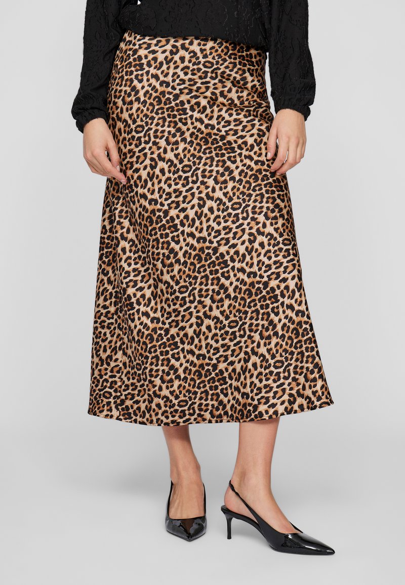 Nő leopárdmintás midi szoknyában, fekete hosszú ujjú blúzban és fekete lakk slingback sarkú cipőben, egy szürke háttér előtt áll.