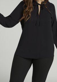 Svart blus med knytkrage och pärlkant, långa ärmar, slät tyg och avslappnad passform. Matchad med texturerade svarta leggings.