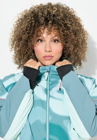 Tyrkysová větrovka s vysokým límcem, černými manžetami a kontrastním prošíváním. Tkanina vypadá hladce s lesklým povrchem. Přední zip je viditelný.