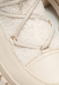 Beżowe obuwie z miękkiego materiału shearling, skórzanymi akcentami i złotymi dodatkami. Model posiada teksturowaną, masywną podeszwę i sznurówki.