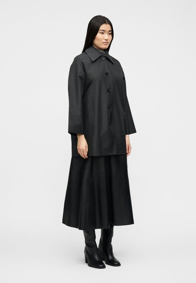 Γυναίκα με μαύρο παλτό με κουμπιά, μακριά μαύρη φούστα και μαύρες μπότες με τακούνι, στέκεται μπροστά σε λευκό απλό φόντο.