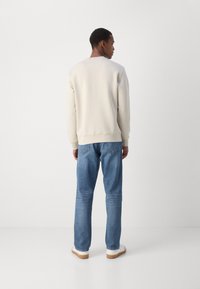 Sudadera beige clara con textura suave, cuello redondo y puños de canalé, combinada con jeans azules. Zapatillas blancas con suelas de goma.
