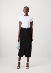 Jupe midi en denim noir avec une fente devant, portée avec un t-shirt blanc moulant. Accessoirisée avec un petit sac à main bleu clair et des talons à lanières.