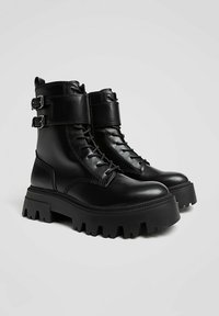 Črni usnjeni bojevniki z debelimi gumijastimi podplati, z zavezovanjem z vezalkami in dvojno sponko na gležnju za nastavljivo prileganje.