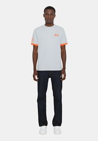 T-shirt grigio chiaro con accenti arancioni sulle maniche, caratterizzato da un logo. Indossato con jeans scuri e sneakers bianche. Vestibilità casual, maniche corte.