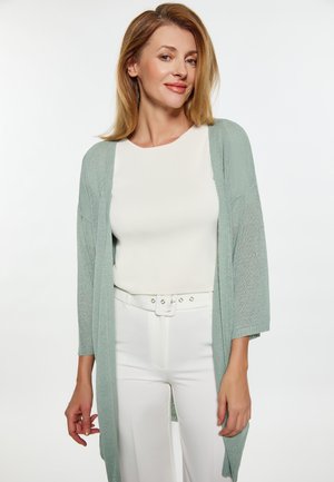 Vrouw met schouderlang blond haar die een lichtgroene cardigan draagt over een wit topje en een witte broek met riem, licht glimlachend.