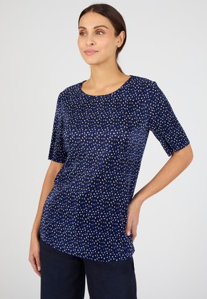 Mujer con un top de manga corta azul marino con pequeños puntos abstractos blancos y azules, combinado con pantalones oscuros, de pie con la mano en la cadera.