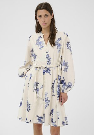 Žena s dlhými hnedými vlasmi v krémových šatách s modrým kvetinovým vzorom, s gombíkmi na prednej strane, viazaním v páse a dlhými nadýchanými rukávmi.