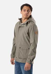 Mladić u svijetlosivoj jakni s kapuljačom i velikim prednjim džepovima te u plavim trapericama, gleda prema lijevo na jednobojnoj pozadini.