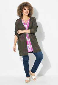 Cardigan aperto di colore verde oliva con due tasche frontali, abbinato a una tunica tie-dye rosa e bianca, jeans blu scuro e sandali bianchi.