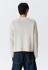 Világos bézs kötött pulóver laza fazonnal, ejtett vállakkal és bordázott nyakkal. Sötét farmernadrággal és látható kulcstartó részlettel párosítva.