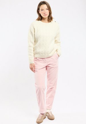 Jeune femme debout, une main dans la poche, portant un pull en maille torsadée blanc, un pantalon rose clair et des baskets beiges sur fond blanc.
