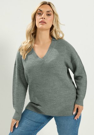 Kvinde med langt blondt hår iført en løs grå strikket V-hals sweater og blå jeans, der står mod en ensfarvet lys baggrund.
