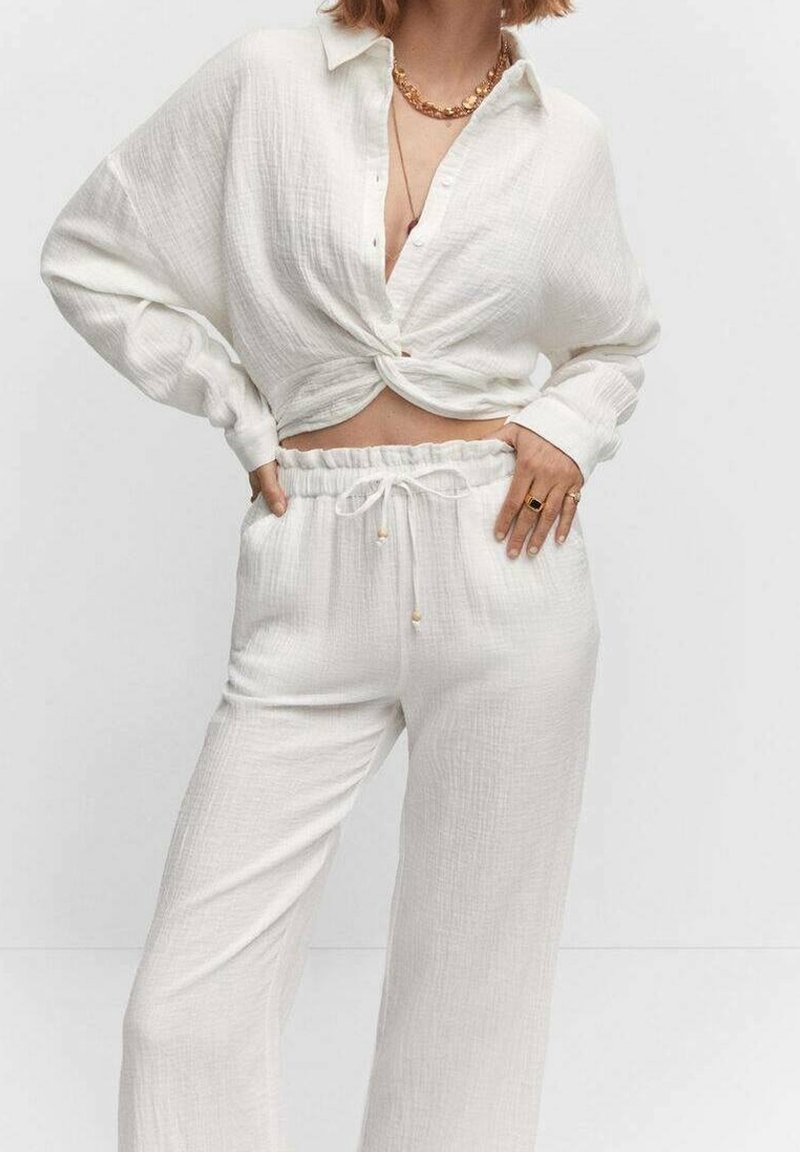 Witte, textuurvolle blouse met een geknoopte voorkant, gecombineerd met losse, met een trekkoord verstelbare broek van dezelfde stof. Gouden sieraden als accenten.