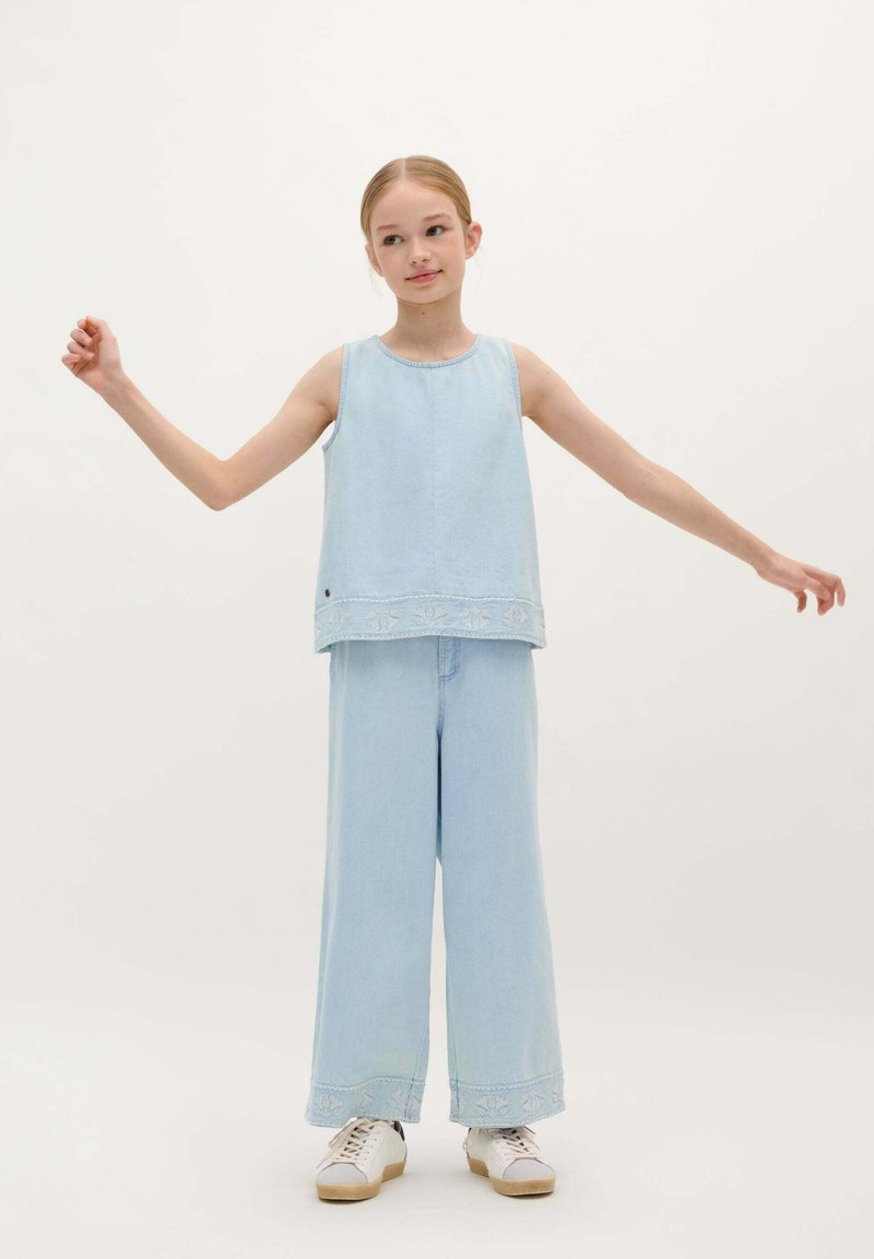 Lichtblauwe denim twee-delige outfit met een mouwloos topje met geborduurde zoom en wijde broek. De schoenen zijn wit met een gestructureerde zool.