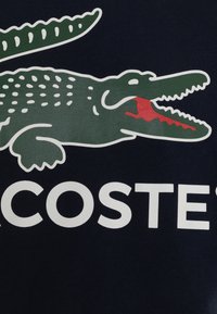 Námorně modré bavlněné tričko s grafikou zeleného krokodýla s bílými akcenty a červeným detailem, doplněné bílým logem "LACOSTE".