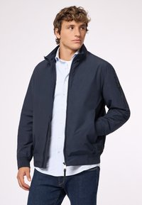 Giacca navy con zip frontale, colletto rialzato e tasche laterali, caratterizzata da una texture liscia e polsini a costine. Abbinata a una camicia a righe leggera.