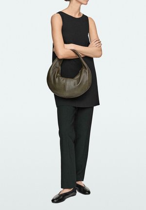 Donna con top nero senza maniche e pantaloni neri, che tiene una grande borsa a mezzaluna di pelle verde scuro con le braccia incrociate.