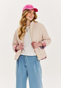 Svetloružová prešívaná bomber bunda so zipsom, vzorovanou podšívkou a zvinutými manžetami. Skombinovaná s bielym tričkom a svetlomodrými džínsami.