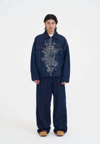 Džínová bunda s výšivkou draka, tmavě modrá barva, zapínání na knoflíky a náprsní kapsy; spárována s volnými džínovými kalhotami a dřeváky.