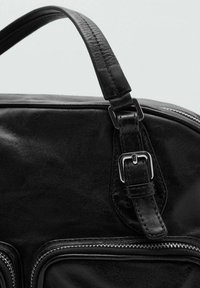 Bolso de cuero negro con textura suave, asa corta con hebilla plateada y cierre de cremallera en los bolsillos laterales.